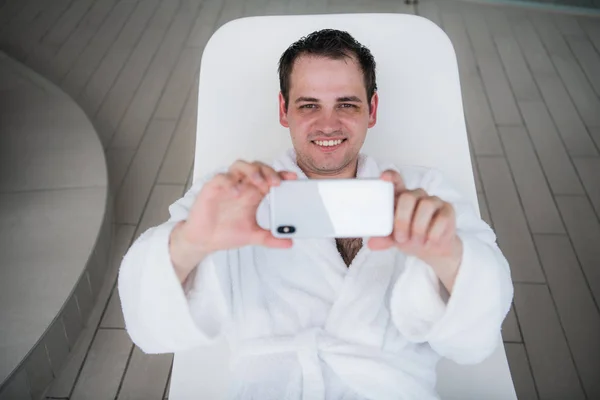 Un hombre con una bata de baño blanca se encuentra junto a la piscina en una silla de cubierta y mira en el nuevo teléfono móvil caro y tomar fotos. El concepto de unas vacaciones de lujo —  Fotos de Stock
