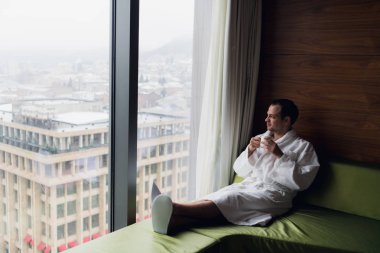 Genç adam akşam kahvesinin ve şehrin güzel günbatımının keyfini çıkarırken pencerenin yanında duruyordu.