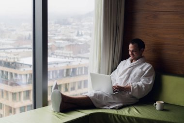 Gündoğumu şehir manzaraya bakarak kahve penceresiyle yakınındaki beyaz banyo bornoz oturma giyen dizüstü bilgisayar üzerinde çalışan genç gülümseyen iş adamı. Motivasyon kavramı