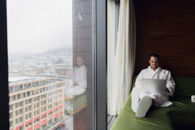 Çalışma evde ya da gezi işadamı. Yan görünümü yatağın üstüne oturan ve dizüstü bilgisayar kullanarak yakışıklı genç işadamı portresi. Dawn güzel şehir manzara arka planda ile panoramik pencere