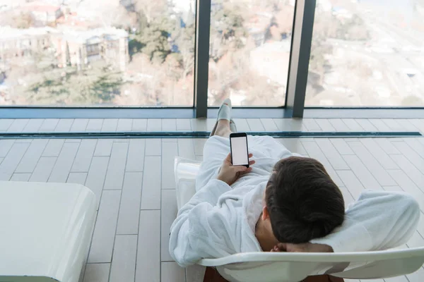 Un joven tumbado en una tumbona cerca de la piscina con teléfono en el interior. Hotel de lujo con una gran vista de la ciudad detrás de la ventana — Foto de Stock