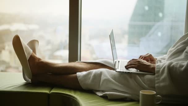 Бізнесмен, який працює вдома або в поїздці. Портрет красивого молодого бізнесмена, який сидить на ліжку і використовує ноутбук. Панорамне вікно з красивими пейзажами міста на фоні — стокове відео