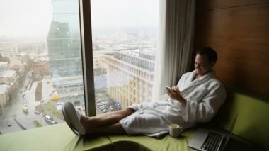 Oturan beyaz banyo bornoz giymiş yakışıklı genç işadamı portresi dizüstü bilgisayar ve cep telefonu gündoğumu city ile modern odasında yatağın arka planda görüntüleyin. Kapalı