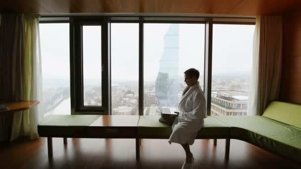 Empresario que trabaja en casa o de viaje. Retrato de vista lateral de joven hombre de negocios guapo sentado en la cama y usando el ordenador portátil. Ventana panorámica con hermosos paisajes de la ciudad del amanecer en el fondo — Vídeos de Stock