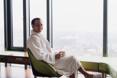 Günaydın dışında bakarken bir fincan kahve zevk modern tam uzunlukta pencere yakınındaki bornoz giyen genç yakışıklı kaygısız adam. Yatay fotoğraf banner Web sitesi header tasarımı ile kopya için