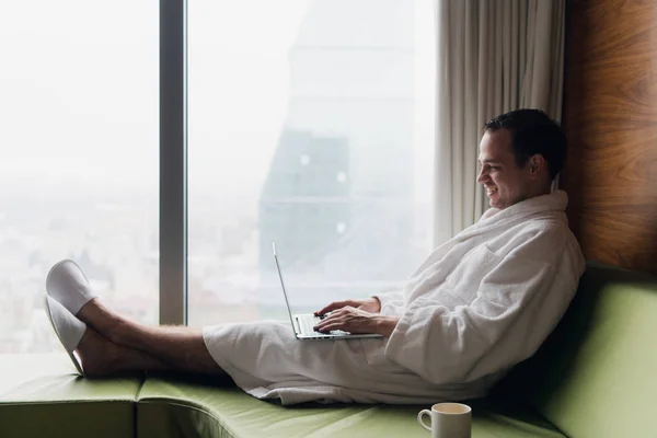 Joven hombre de negocios sonriente que trabaja en el ordenador portátil con bata de baño blanco sentado cerca de la ventana con taza de café mirando al amanecer vista de la ciudad. Concepto de motivación —  Fotos de Stock
