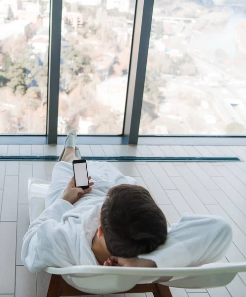 Un joven tumbado en una tumbona cerca de la piscina con teléfono en el interior. Hotel de lujo con una gran vista de la ciudad detrás de la ventana — Foto de Stock
