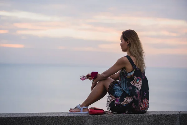 Красивая женщина сидит на набережной и наслаждается видом, в коротком платье, с мобильного телефона — стоковое фото