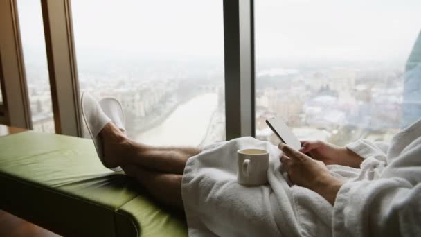 Empresario que trabaja en casa o de viaje. Retrato de vista lateral de joven hombre de negocios guapo sentado en la cama y usando el ordenador portátil. Ventana panorámica con hermosos paisajes de la ciudad del amanecer en el fondo — Vídeos de Stock