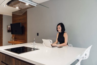 Güzel iş kadını dizüstü bilgisayar kullanıyor, kameraya bakıyor ve evde otururken gülümsüyor.