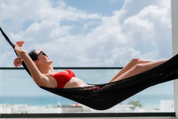 Back view of woman in red bikini resting in hammock with ocean view on rooftop of a luxury hotel.