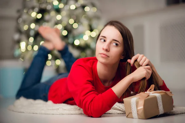 Fiatal mosolygós nő pulóverben kezében egy ajándék doboz ünnepli téli ünnepek díszített otthon belső karácsonyfával. Boldog új évet koncepció — Stock Fotó