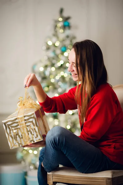 Feiertage, Feiern und Menschen Konzept - lächelnde Frau in rotem Pullover und Jeans hält goldene Geschenkschachtel über Weihnachtsbaumbeleuchtung Hintergrund — Stockfoto