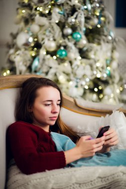 Genç kadın o liyng kanepe önünde evde Noel ağacı olarak cep telefonuna metin mesajlarını kontrol.