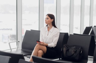 Yolculuğuna başladı. Güzel genç bir kadın, havaalanı lobisinde uçağa binmeyi beklerken pencereden uçan uçağa bakıyor..