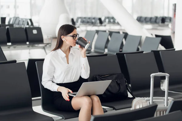 Young female managing director is reading e-mail on laptop and listening music, while is waiting her fly in international airport hall. Woman lawyer with take away coffee is watching financial news on — Stock Photo, Image