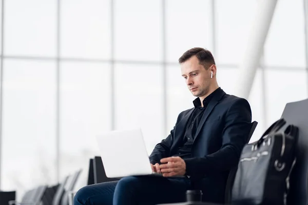 Mann mit Koffer sitzt im Flughafen-Wartebereich und hört Musik mit Airpods — Stockfoto