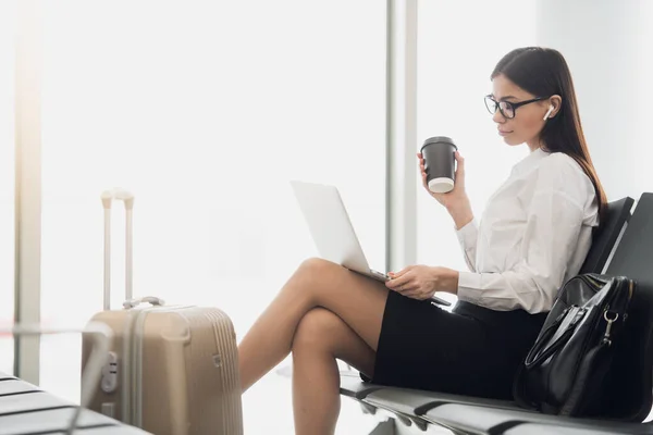 La joven directora gerente está leyendo e-mail en el portátil y escuchando música, mientras espera su vuelo en la sala del aeropuerto internacional. Una abogada con café para llevar está viendo noticias financieras en —  Fotos de Stock