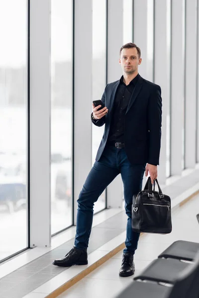 Geschäftsmann mit Handy-App am Flughafen. Junger Geschäftsmann, der mit dem Smartphone durch Bürogebäude oder Flughafenterminal läuft. Schöner Mann mit stylischer Anzugjacke drinnen. — Stockfoto