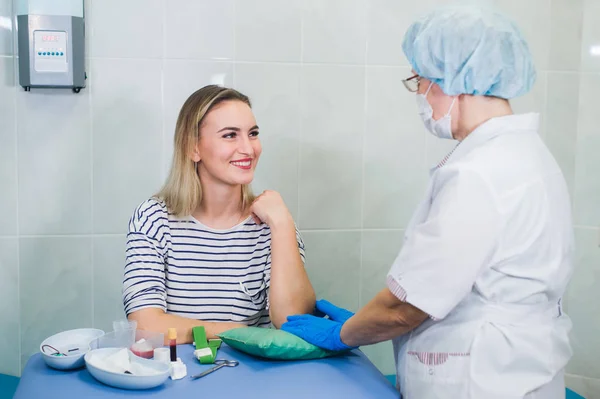 Preparación para el análisis de sangre con una hermosa mujer rubia joven por parte de una doctora en uniforme médico de bata blanca en la mesa en una habitación blanca brillante. Enfermera frotando un tejido estéril de la mano del paciente . — Foto de Stock