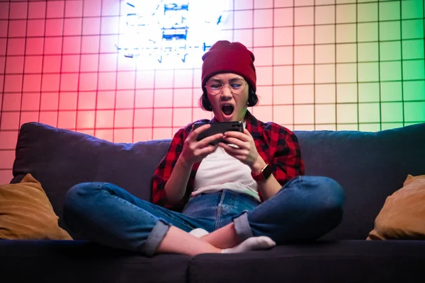 Nahaufnahme Porträt von hübschen Teenager-Mädchen beim Online-Spiel in Telefon trägt lässige Jeans Jeanshemd Outfit Kleidung sitzt auf einem gemütlichen Sofa — Stockfoto