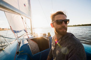 Serious handsome young male yacht captain in sunglasses wearing gloves staying on boat deck and looking into distance