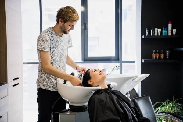 Conceito de beleza e pessoas - mulher jovem feliz com cabeleireiro cabeça de lavagem no salão de cabeleireiro — Fotografia de Stock