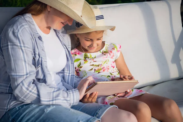 Mãe feliz e sua filhinha desfrutando de tempo livre usando o computador tablet enquanto relaxa no jardim da casa de férias durante o dia ensolarado . — Fotografia de Stock