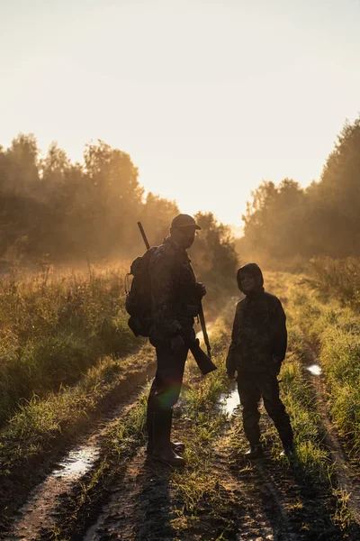 Hunters with hunting equipment going away through rural field towards forest at sunset during hunting season in countryside. — Stock Photo, Image