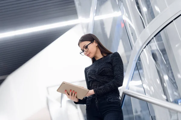 Young serious beautiful female business woman standing on the stairs and using tablet while waiting for colleague.