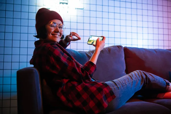 MOSCÚ, RUSIA - Diciembre 2019: vista trasera de un primer plano de una mujer jugando juego en línea en un teléfono inteligente tumbado en un sofá en casa — Foto de Stock