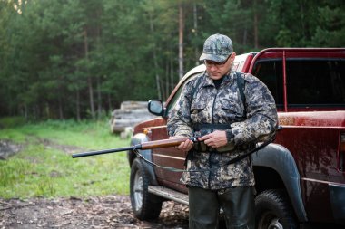 Avcı erkek silah taşıyor, günlük av kıyafetleri giyiyor, sırtına bıçak ve yiyecek koyuyor. Orman arkaplanı.