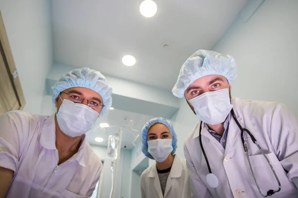 Γιατροί με ιικές προστατευτικές άσπρες μάσκες που δουλεύουν σε επείγουσα βοήθεια κοιτούν την κάμερα από ψηλά όπως εσύ ως ασθενής. Προσπαθούν να σταματήσουν την επιδημία του στέμματος του ιού.. — Φωτογραφία Αρχείου