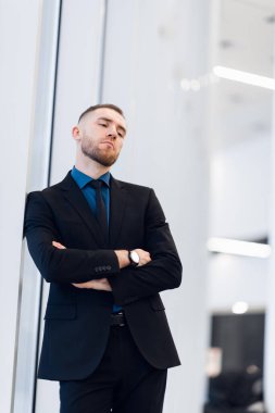 Orta yaşlı bir adam, kollarını kavuşturmuş, pencerenin kenarına yaslanmış kameraya bakıp düşünceli bir ifadeyle bakıyor.