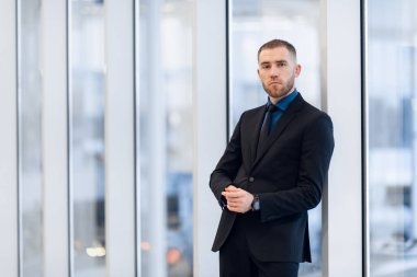 Orta yaşlı bir adam, kollarını kavuşturmuş, pencerenin kenarına yaslanmış kameraya bakıp düşünceli bir ifadeyle bakıyor.