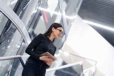 Genç, ciddi, güzel bir iş kadını merdivenlerde duruyor ve meslektaşını beklerken tablet kullanıyor..