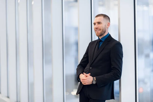 Stylish young businessman wearing a modern suit, who is a high achiever, standing on the top floor of an office building looking out at the view through large windows — Stock Photo, Image