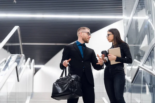 Biznesowa para na schodach rozmawiająca i uśmiechnięta. Mężczyzna w garniturze i okularach z torbą rozmawiający z kobietą trzymającą tablet. — Zdjęcie stockowe