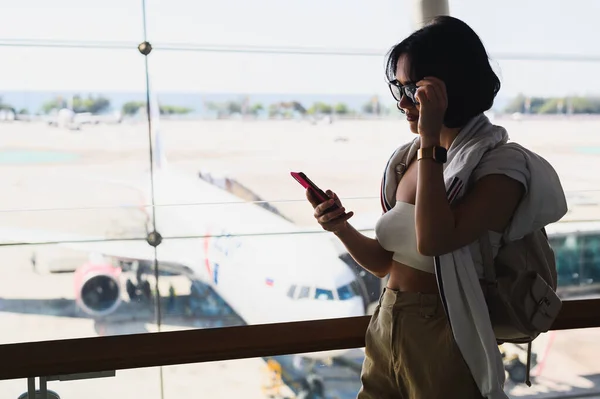 Mujer joven está navegando en Internet por teléfono móvil en la sala del aeropuerto o estación. Ella está mirando en la exhibición de gadget y deslizando por el dedo —  Fotos de Stock