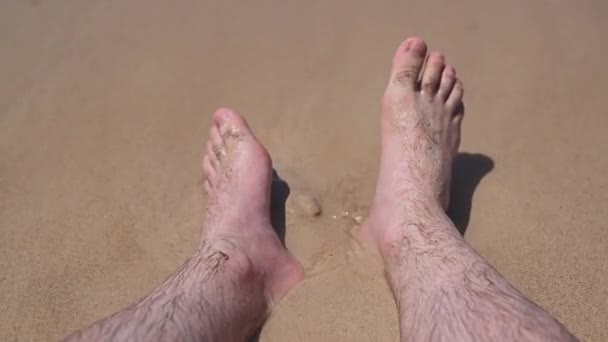 Mans láb mosta óceán hullámok és eltemeti őket a homokba. Nézőpont a fiatal férfi ül az arany homok a tengerparton. — Stock videók