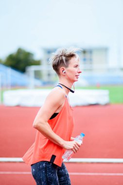 Genç bir kadın müzik dinleyerek ve gülümseyerek stadyuma koşuyor. Bir şişe su tutuyorum.