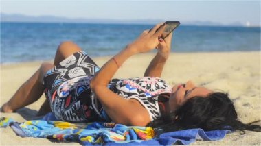 Güzel bir esmer güzel bir kadın kumlu bir plaj üzerinde yalan, sahilde yazma, telefonda mesaj yazmak ve şehvetli bir gülümseme ile gülüyor. Düşük bir açıdan güneşli bir günde vurdu.