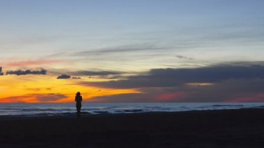 Mutlu gün batımında plaj boyunca yürüyen genç ve güzel kadın. Uzaktan statik kayıt ve kadın tarafından çekilen video zevk ve mutluluk Karadağ'ın kumlu Sahili güzel sahne gösterir.