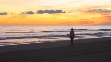 Mutlu gün batımında plaj boyunca yürüyen genç ve güzel kadın. Statik bir resim ve kadın ile uzaktan alınan bir video kameradan yavaş hareket ve Karadağ'ın kumlu Sahili güzel sahne zevk ve mutluluk gösterir.
