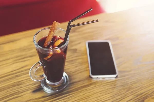 Glühweinglas und Mobiltelefon auf Tischplatte Hintergrund verschwimmen. Rotwein mit Zimt, Nelken, Anis, Zitrusfrüchten zum Aufwärmen im Café. süße, würzige und wärmende Wintergetränke. Gluhweintrinken allein — Stockfoto