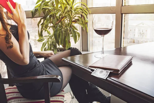 Joven mujer bonita sentada en el restaurante bebiendo una copa de vino mirando por la ventana panorámica con una hermosa vista de la ciudad y hablando por teléfono. 100 dólares en la carpeta para la cuenta en la mesa . — Foto de Stock