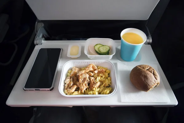 Varios alimentos de clase económica y un teléfono inteligente de pasajeros con una pantalla en blanco en la mesa plegable del avión. Comidas a bordo del avión. Cuchillería de plástico cuchillo y tenedor en un plato con comida . — Foto de Stock