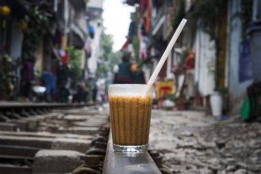 a glass of brown cold coffee is on the railway in Hanoi. Vietnam. One of the attractions of Hanoi - a train passing between the old houses on a narrow city street. clipart