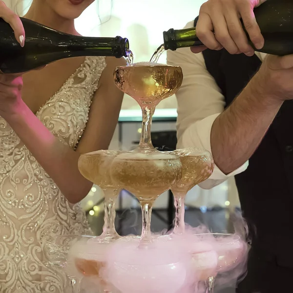 Close up of a cascade champagne fountain made by groom and bride at banquet background. Newly married pour sparkling wine into the glasses tower. Filling glasses with champagne at wedding celebration