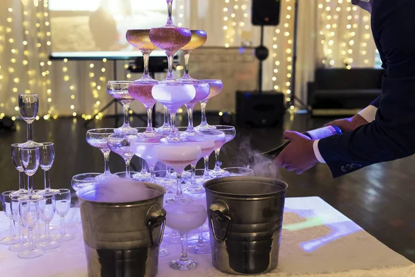 ライトアップされた宴会場の背景には、グラスタワーにシャンパンが注がれます。スパークリングワインのグラスピラミッドは、宴会テーブルの上のウェイターによって作られています。お祝い事に出す — ストック写真